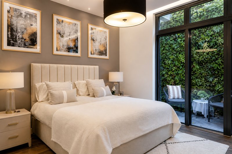 Modern bedroom with cream upholstered bed, bedside lamps, framed wall art, and French doors opening to garden terrace