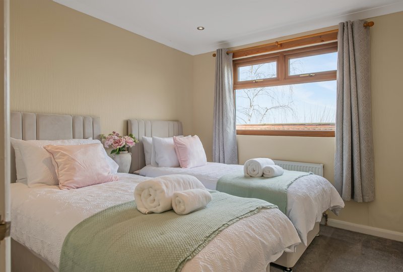 Bright bedroom with two single beds, neutral bedding, pink cushions, rolled towels, and large windows.
