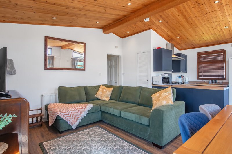 Open-plan living room with green corner sofa, wooden ceiling, kitchenette, and hardwood floors.