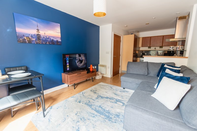 Modern open-plan living room with blue accent wall, gray sofa, wooden TV stand and kitchen area