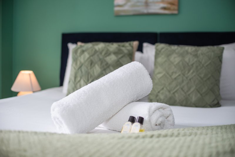 Bedroom with green accent wall, black bed frame, white pillows and rolled towel with toiletries