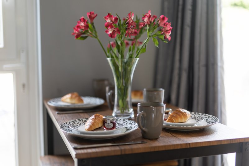 Dining table set with pink flowers, croissants, plates and glasses by window