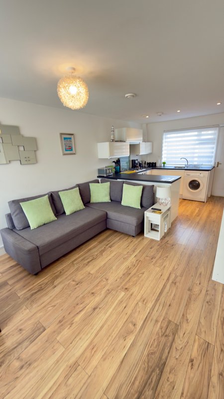 Open-plan living room with grey corner sofa, wooden flooring, kitchen area, and pendant lighting
