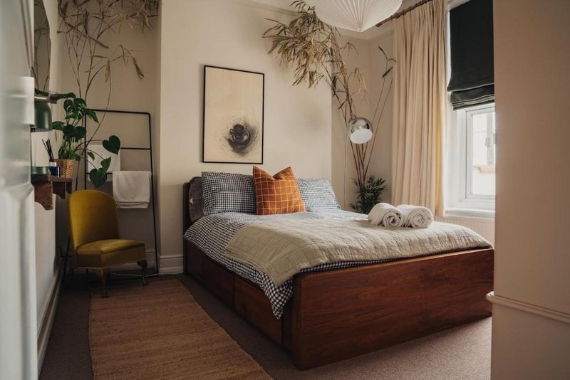 Bedroom with wooden bed frame, neutral bedding, plants, framed wall art, and mustard armchair