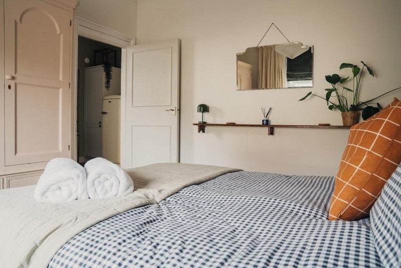 Bedroom with double bed, blue gingham bedding, white pillows, orange cushion and wall shelf