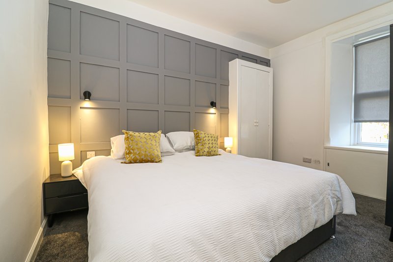 Modern bedroom with double bed, grey panelled headboard, yellow cushions and bedside lighting