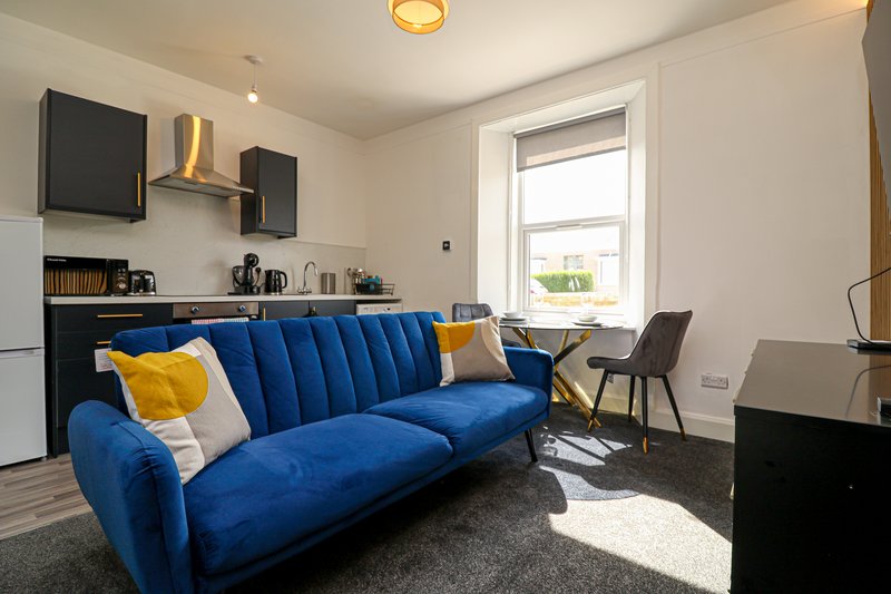 Modern living room with blue velvet sofa, yellow cushions, kitchenette, and natural light from window