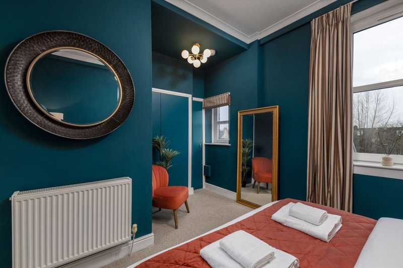 Modern bedroom with teal walls, double bed with red throw, round mirror, radiator and large window