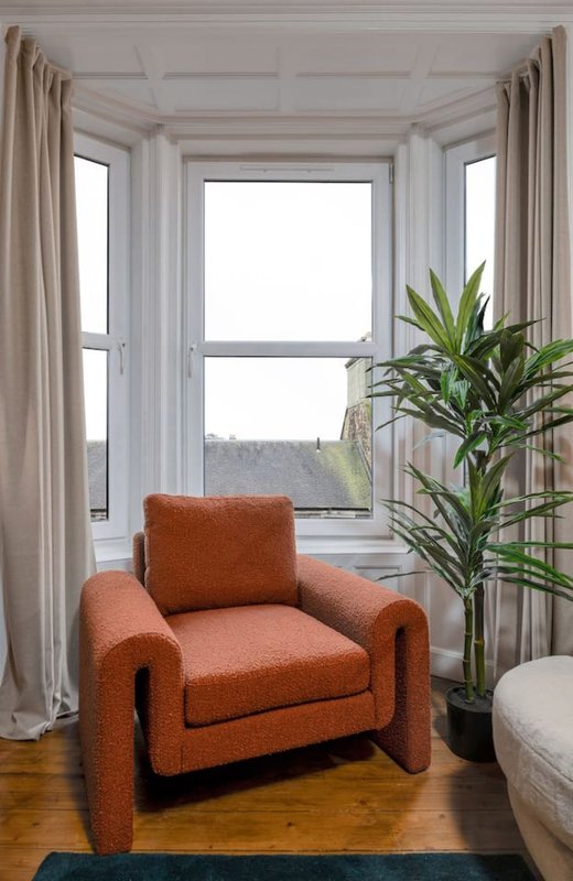 Bright sitting area with orange armchair, large bay window, cream curtains and potted plant