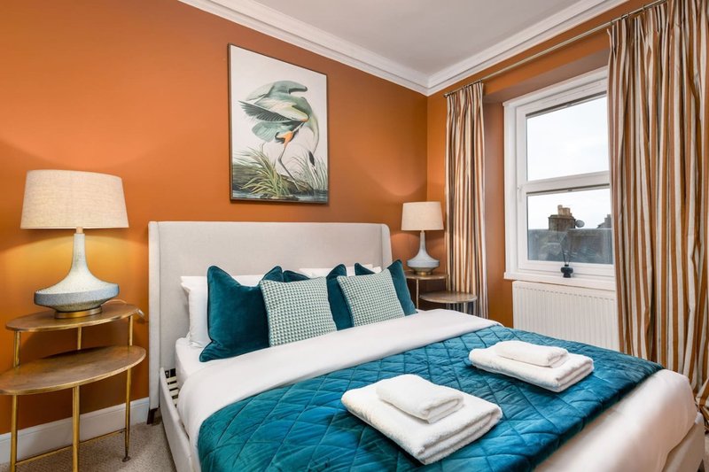 Bright bedroom with orange walls, double bed, teal bedding, white pillows and window with curtains