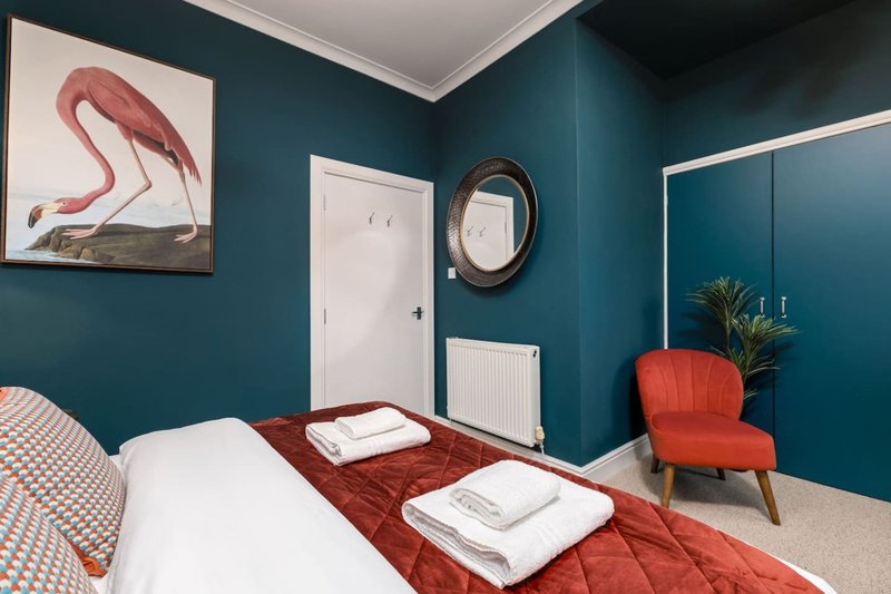 Bedroom with teal walls, double bed, red accent chair, framed flamingo artwork and mirror