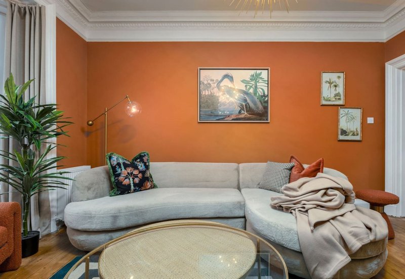 Modern living room with curved gray sofa, orange walls, framed artwork, and wooden flooring