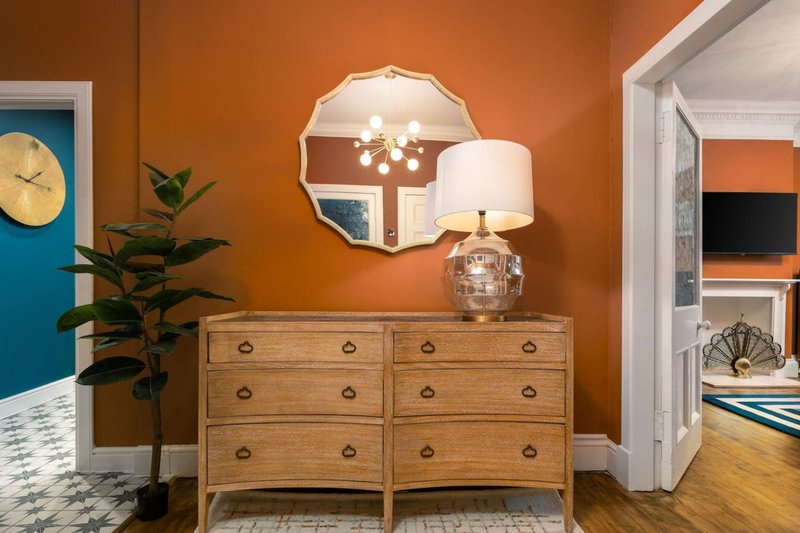 Bedroom corner with wooden dresser, decorative mirror, table lamp and potted plant against orange wall