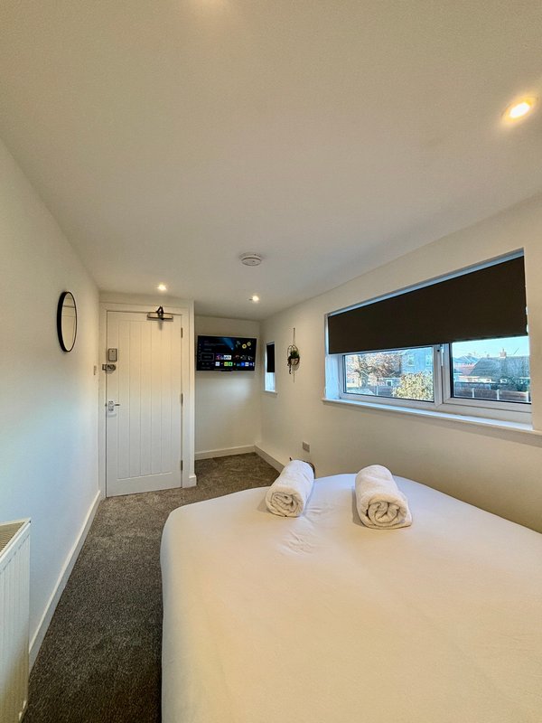 Bright bedroom with white bed, rolled towels, window with dark blinds, white door and recessed lighting