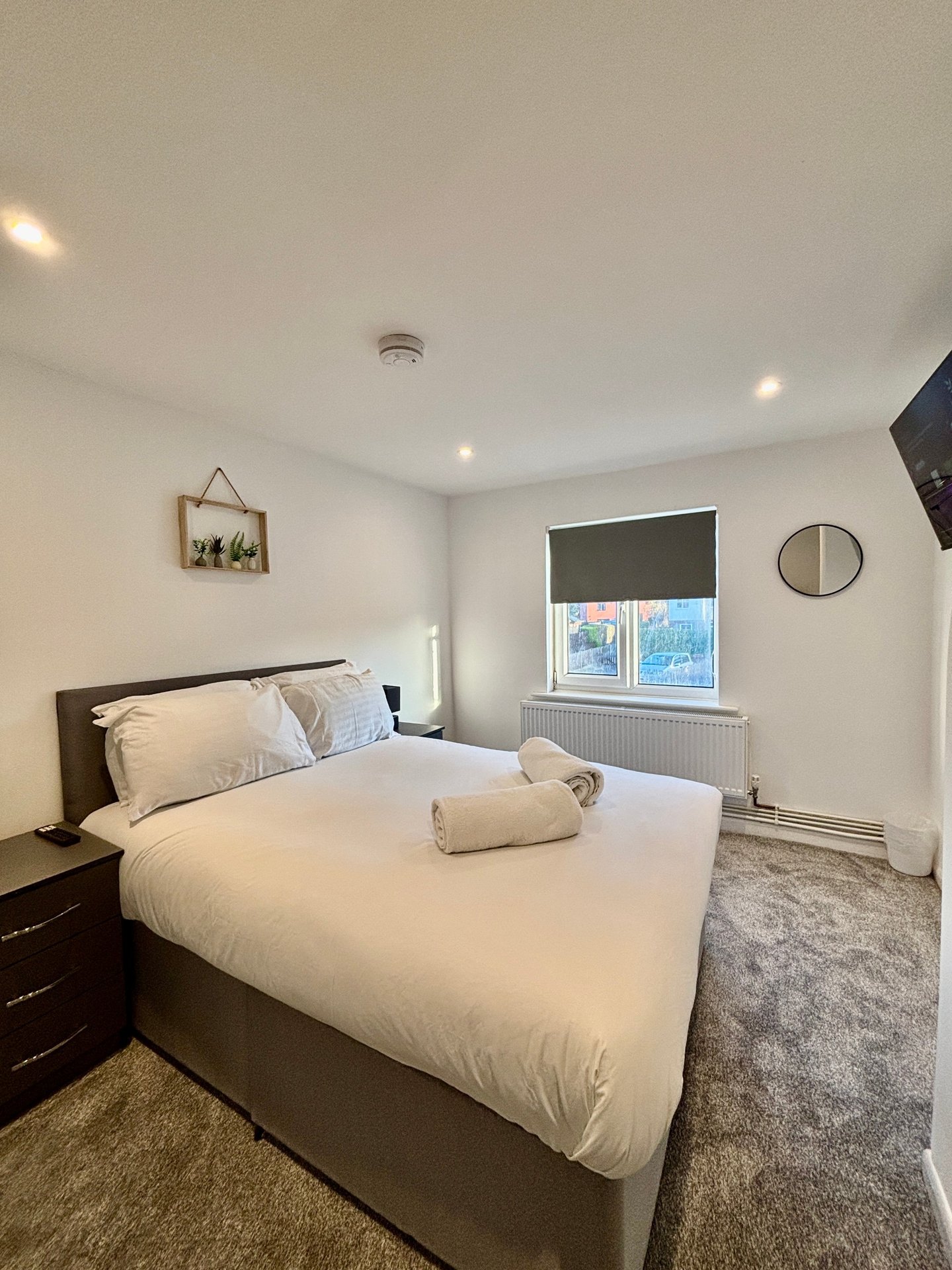 Bright bedroom with double bed, white bedding, dark wood nightstand, round mirror and window with blind
