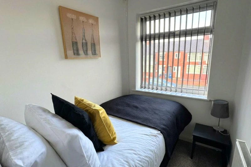 Bedroom with double bed, black and gold cushions, bedside table with lamp and window with vertical blinds
