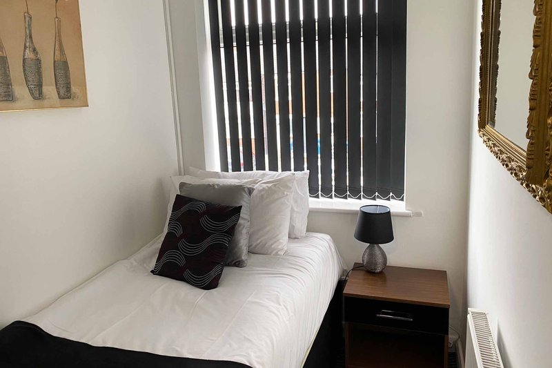 Small bedroom with single bed, white bedding, black cushion, wooden bedside table and lamp
