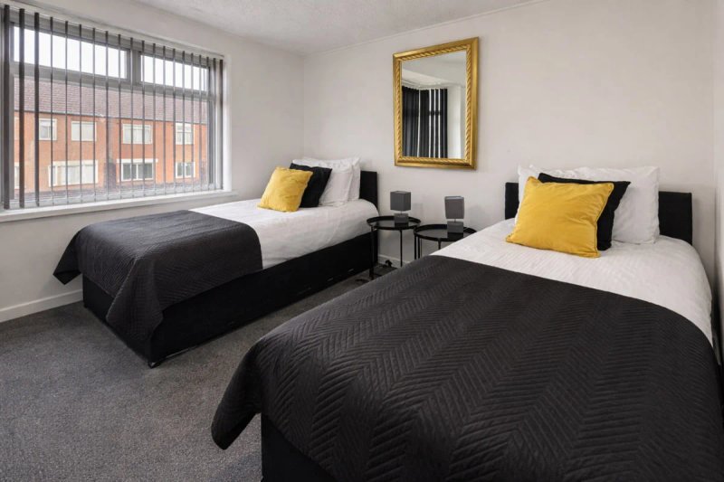 Twin bedroom with two single beds, black bedding, yellow cushions, mirror and window with blinds