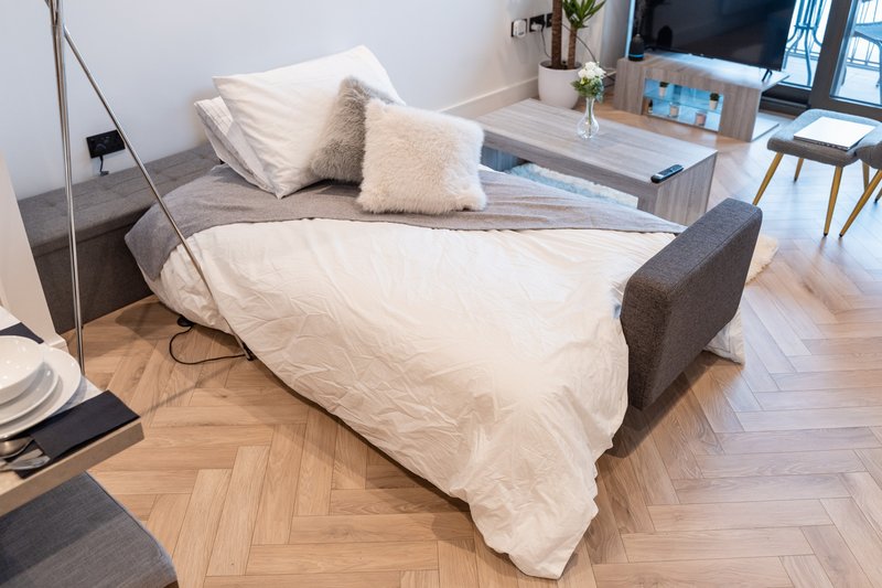 Modern studio bedroom with sofa bed, cream bedding, cushions, wooden floor and TV