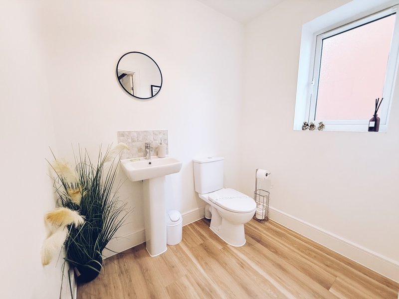 Clean bathroom with toilet, pedestal sink, circular mirror, wooden flooring and small window