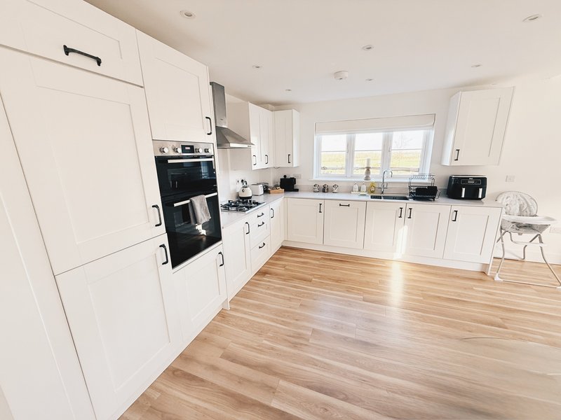 Modern white kitchen with wooden flooring, built-in oven, and large window overlooking garden