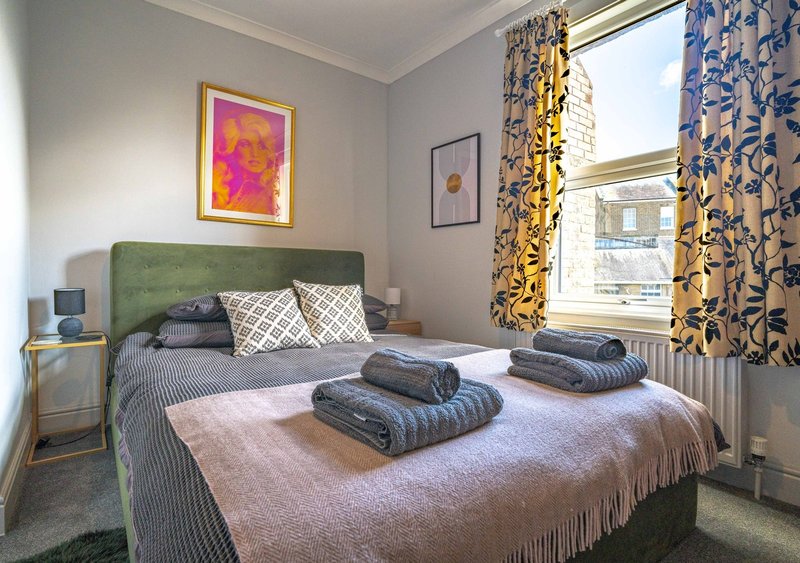 Bedroom with green headboard, double bed, folded towels, framed artwork and patterned curtains