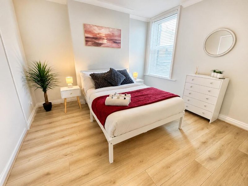 Bright bedroom with white bed, red bedding, wooden floor, nightstands and dresser