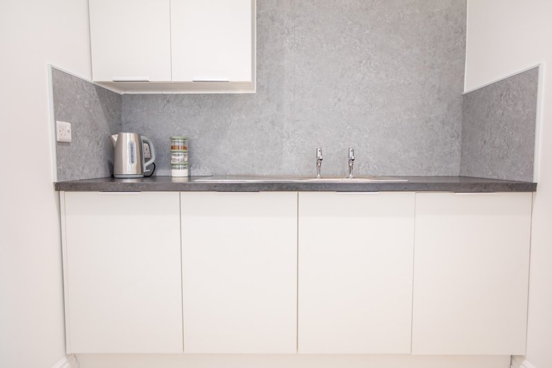 Modern kitchenette with white cabinetry, gray countertop, kettle and jar on shelf