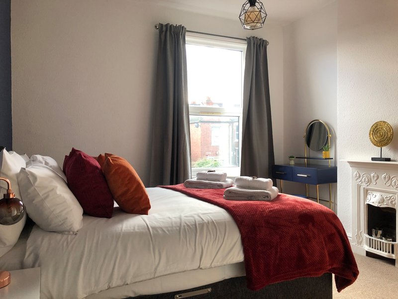 Bedroom with double bed, red throw blanket, decorative pillows, window with grey curtains