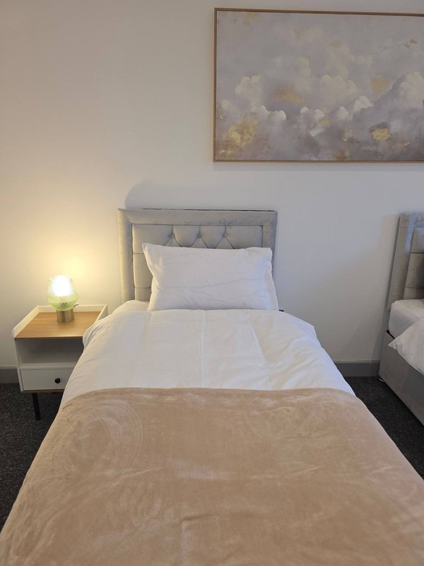 Single bed with grey upholstered headboard, white pillows, beige throw, bedside lamp and nightstand
