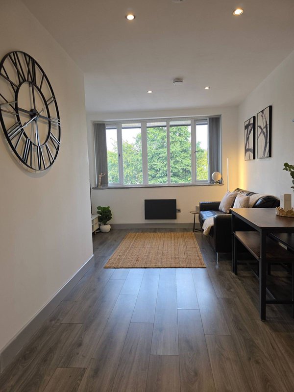 Modern open-plan living area with wooden flooring, large window, wall clock and desk space