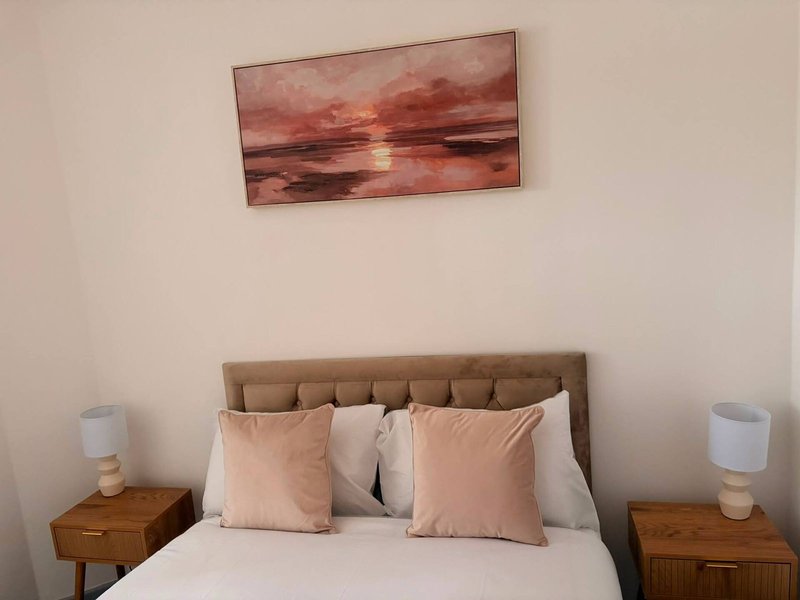 Bedroom with double bed, beige headboard, pink cushions, wooden nightstands and sunset artwork