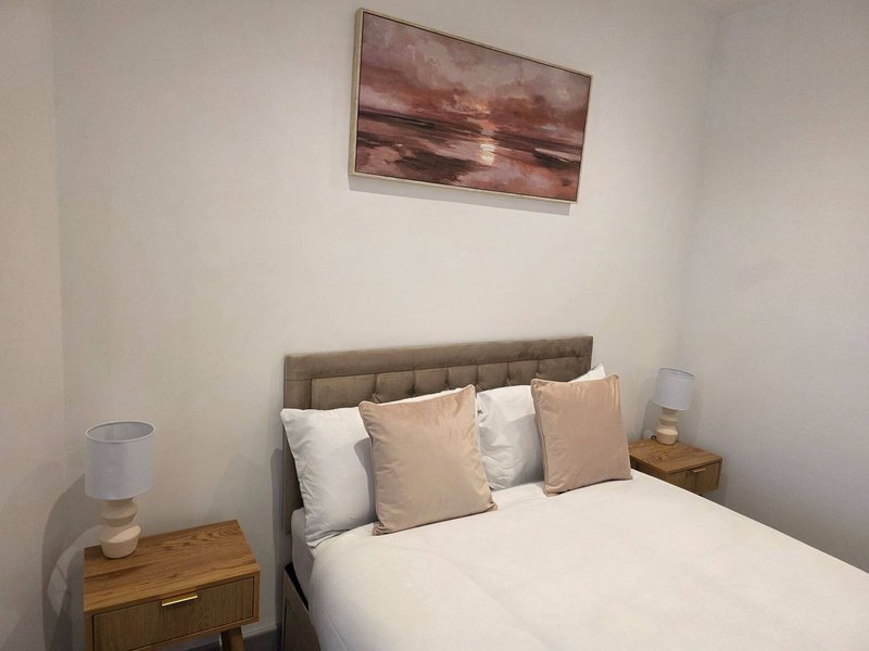 Bedroom with double bed, grey headboard, cream and beige pillows, wooden bedside table, and framed abstract artwork above bed