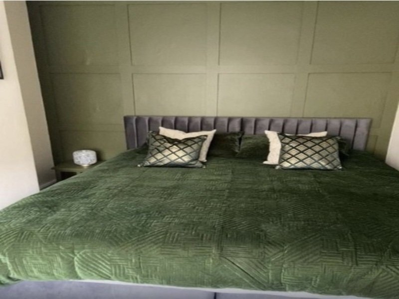 Bedroom with grey upholstered headboard, green quilted bedding, patterned pillows and white bedside lamp
