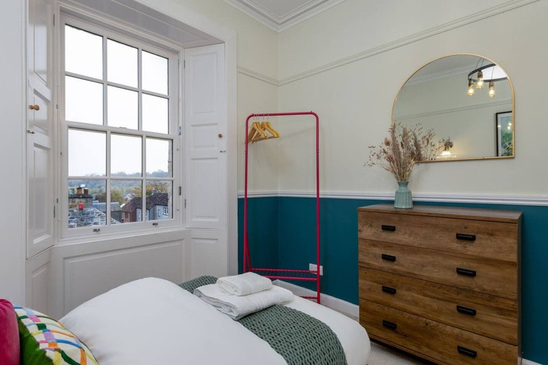 Bedroom with white bed, wooden chest of drawers, round mirror, and large window with countryside view