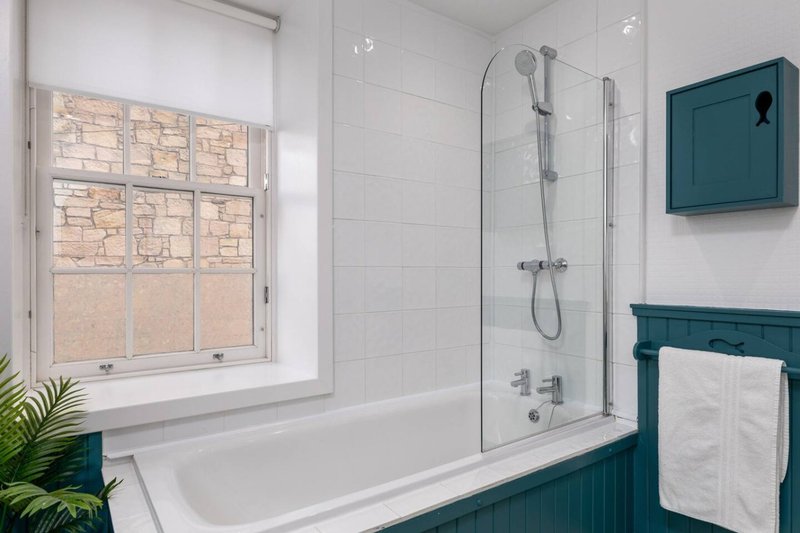 Modern bathroom with white tile walls, teal panelling, bathtub with glass shower screen and window