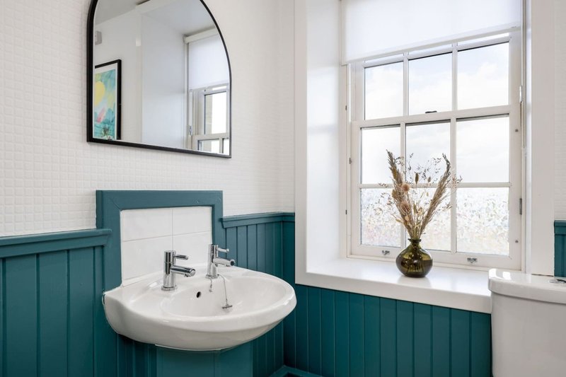 Bathroom with white sink, teal paneling, round mirror and window with dried flowers