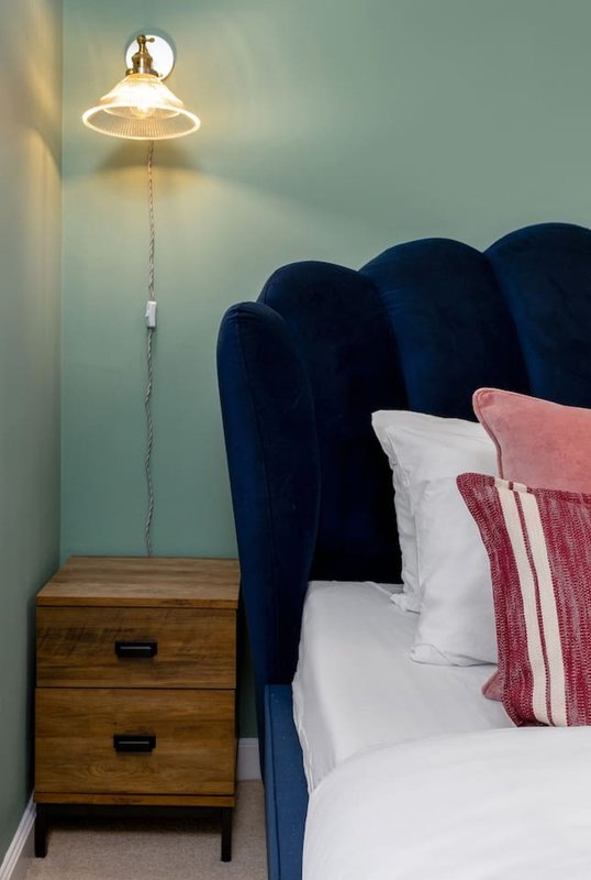 Bedroom corner with blue upholstered headboard, white bedding, burgundy cushions, wooden bedside table, and pendant wall lamp