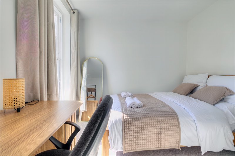 Bright modern bedroom with double bed, wooden desk, chair and large window with curtains