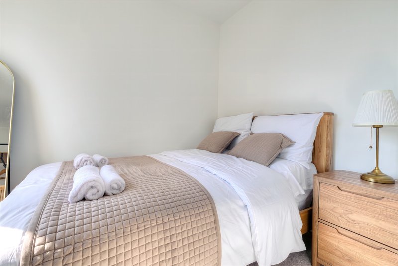 Bright bedroom with wooden bed frame, white bedding, rolled towels, and brass table lamp