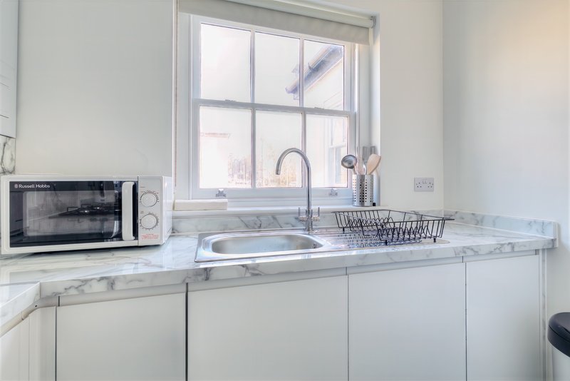 Modern white kitchen with marble countertop, sink, microwave, and window overlooking street