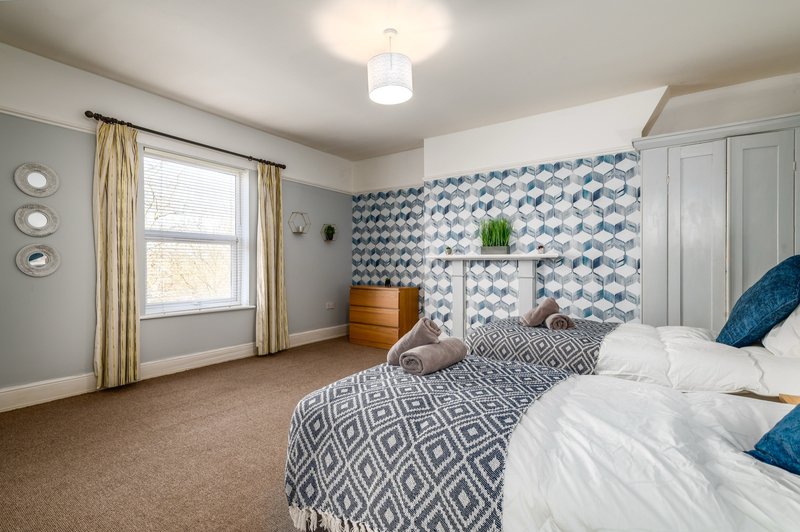 Bright bedroom with two beds, patterned accent wall, wooden dresser, and window with gold curtains