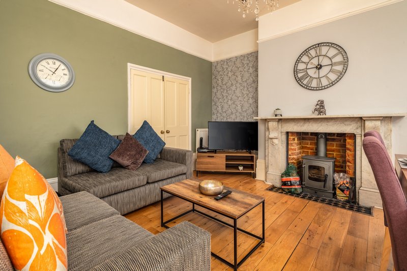 Living room with grey sofa, fireplace with stove, wooden floor and wall clocks