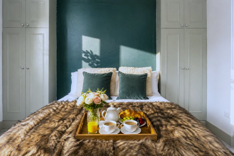 Bedroom with dark green accent wall, double bed, white storage cabinets, decorative tray with flowers and breakfast items
