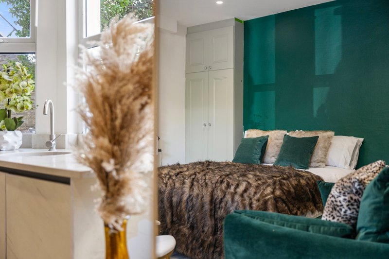 Bedroom with green accent wall, double bed with patterned bedding, and window with kitchen sink visible
