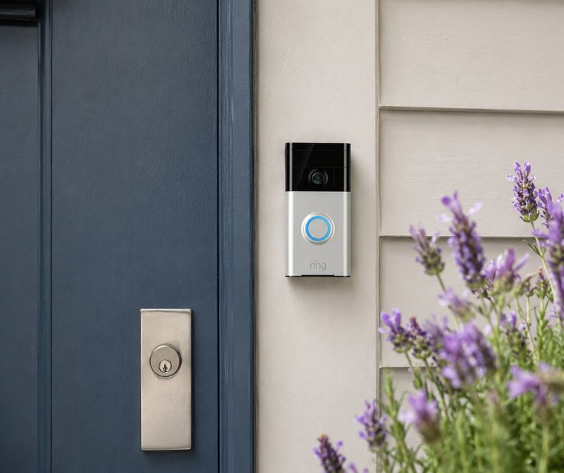 Front door entrance with doorbell camera, lock, and purple flowering plants