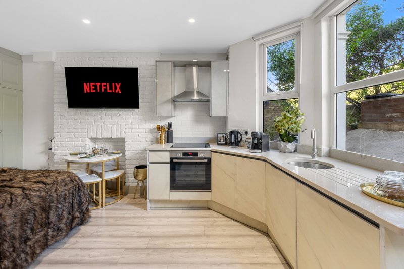 Modern open-plan kitchen and living area with cream cabinets, white brick, TV on wall, and garden views