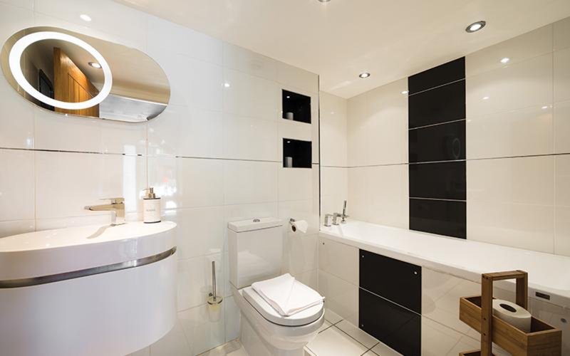 Modern bathroom with white vanity, illuminated mirror, toilet, bathtub and black tile accents