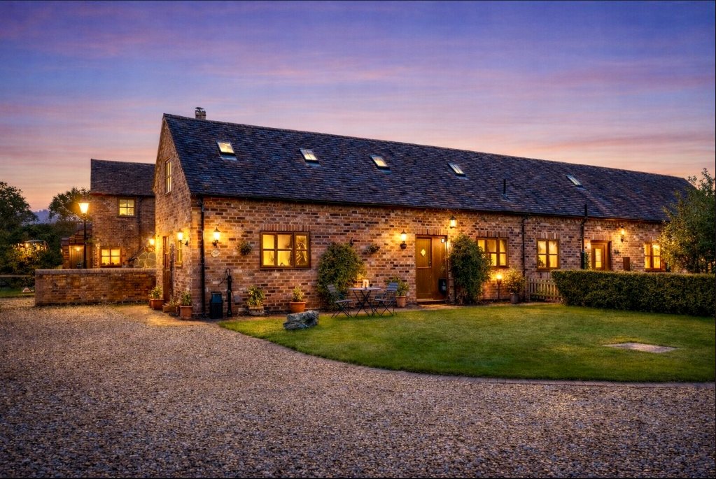Stone cottage exterior at dusk with illuminated windows, gravel driveway and manicured lawn