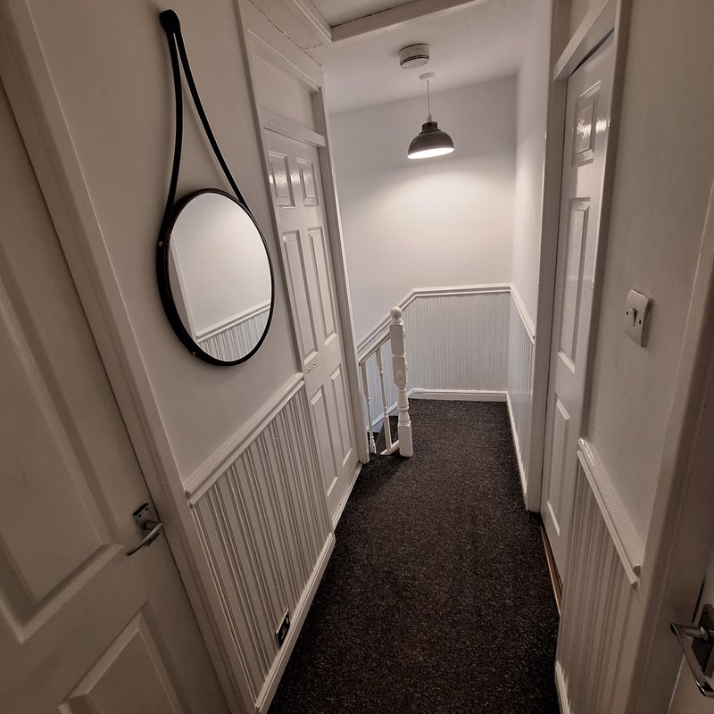 Narrow hallway with white walls, dark carpet, round mirror, pendant light and staircase.